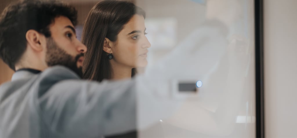 A man and a woman are working together, focusing on a whiteboard or screen in a collaborative environment. The man is pointing at something on the board while the woman observes intently. Both are dressed in professional attire, with the woman wearing earrings and having long, dark hair. The image captures a moment of concentration and teamwork, with both individuals engaged in problem-solving or strategizing. The scene emphasizes collaboration and attention to detail in a professional setting.