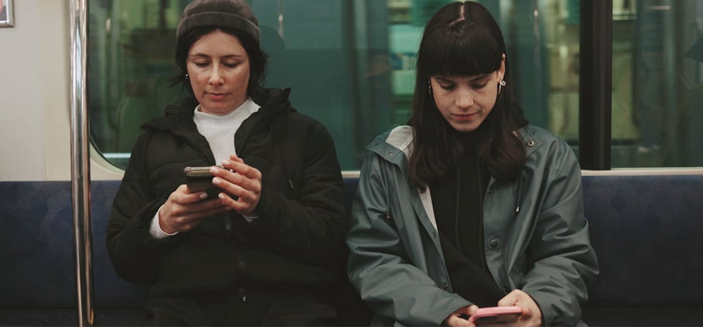 Two women are sitting side by side on public transportation, both focused on their smartphones. The woman on the left, wearing a beanie and a black jacket, is holding her phone with both hands. The woman on the right, in a teal jacket, is also engaged with her phone, looking down at the screen. The atmosphere is quiet and reflective, as both individuals are absorbed in their own devices. The setting is inside a train or subway car, with the city passing by outside the window. The image conveys the modern-day experience of commuting with technology.