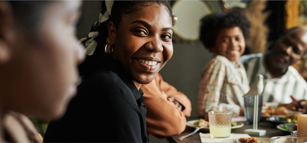 A young woman is smiling warmly at the camera while sitting at a dinner table with her family. She is wearing a black shirt and gold earrings, and her hair is tied back with a patterned scarf. In the background, other family members, including a young boy and an older man, are also smiling and enjoying the meal. The table is set with plates of food, drinks, and a candle, creating a cozy and welcoming atmosphere. The image captures the joy of shared moments and connection during a family gathering.