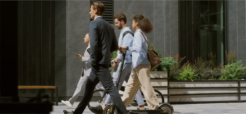 A group of professionals is walking down a city street. One man in a business suit is carrying a helmet and walking with purpose, while another man walks with a folded bike. A woman pushes an electric scooter as she walks, and another woman is checking her phone as she keeps pace with the group. The urban environment is modern, with sleek buildings and greenery lining the walkway. The image captures a mix of personal mobility options and professional life in a bustling city, highlighting sustainable and efficient urban commuting.