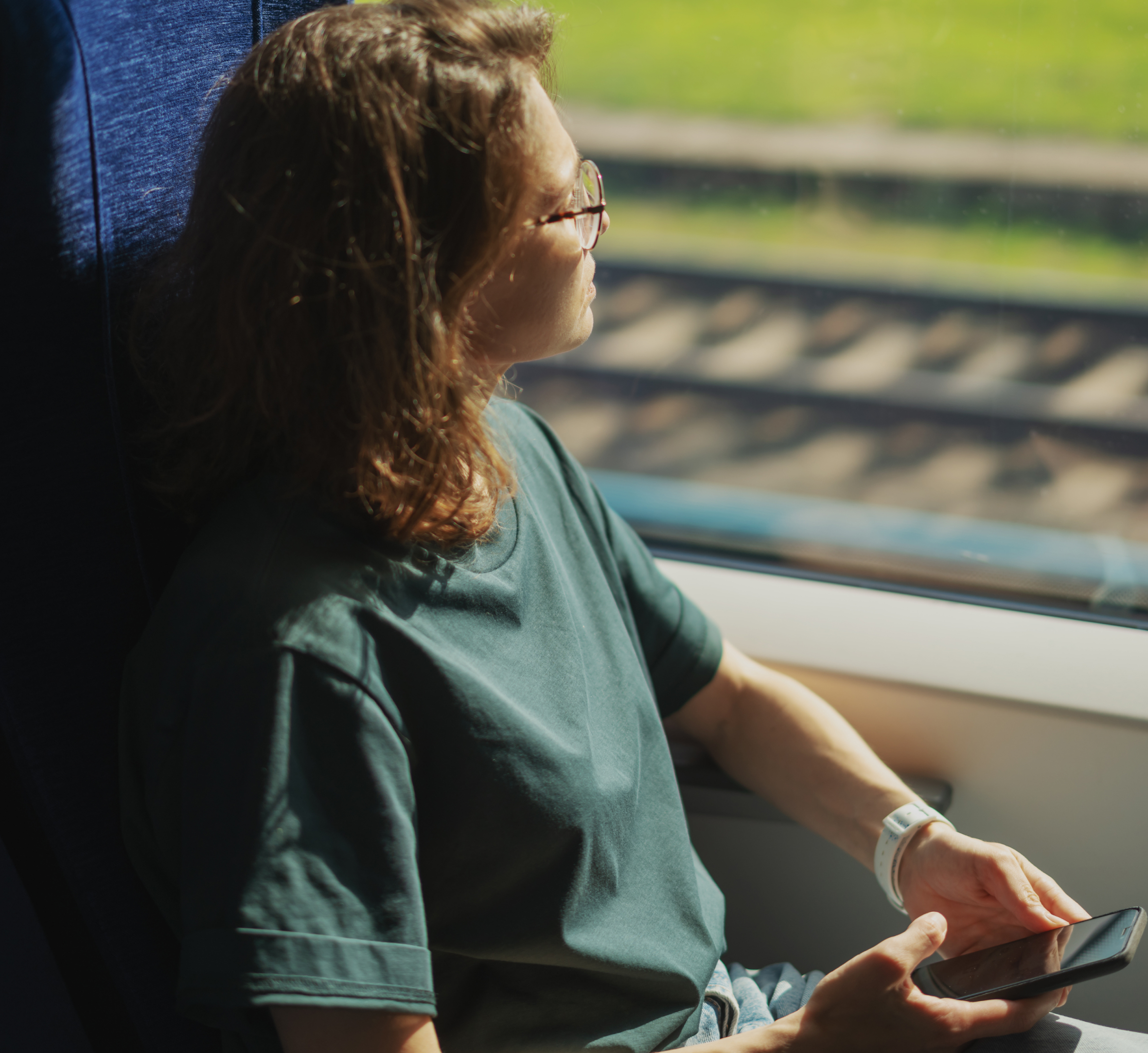 A young woman with shoulder-length hair and glasses is sitting by a train window, gazing out at the passing scenery. She is casually dressed in a green t-shirt and holding her smartphone in one hand, resting it on her lap. The sunlight illuminates her face and the train seat, while the blurred view of railway tracks and greenery outside adds to the feeling of peaceful travel. The image captures a quiet moment of reflection and relaxation during a train journey.