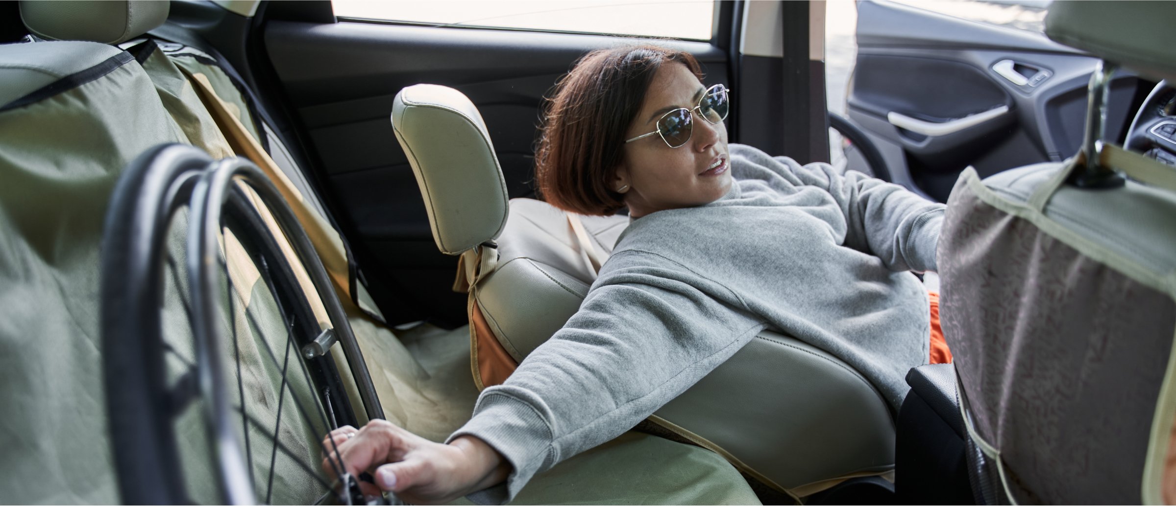 A woman wearing sunglasses is sitting in the back seat of a car, reaching for the wheel of a folded wheelchair next to her. She is dressed casually in a gray sweatshirt and appears relaxed. The car door is open, allowing sunlight to fill the space. The image highlights independence and mobility, showing the woman comfortably managing her wheelchair while traveling. The interior of the car is practical and functional, supporting her needs.